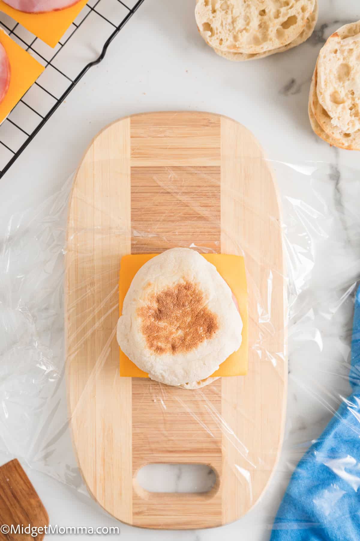 An English muffin half with a slice of cheese sits on a wooden cutting board covered with plastic wrap. Other food items and kitchen utensils are partially visible on a marble countertop.