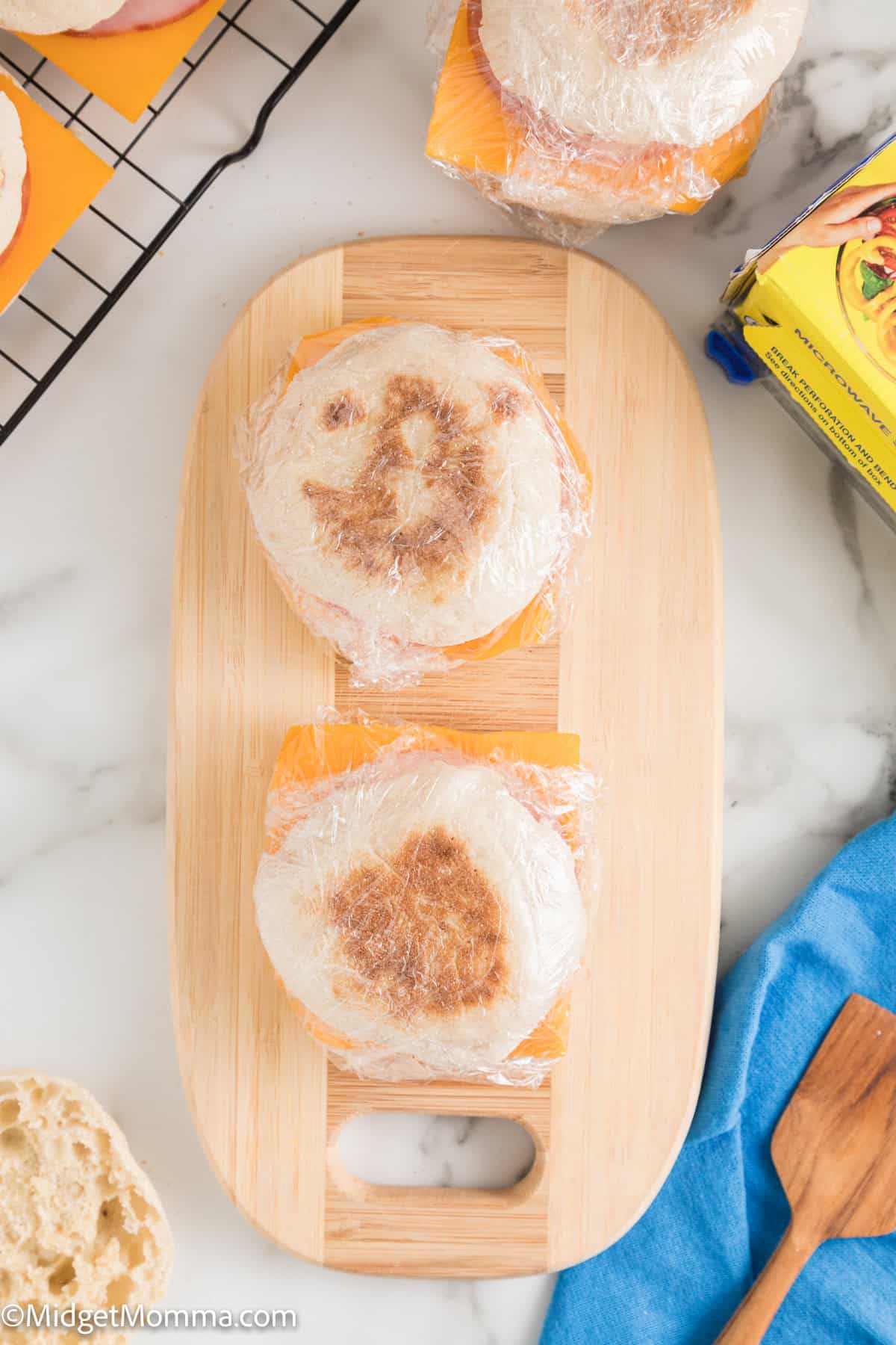 Two wrapped English muffins with cheese slices on a wooden board. One has a pattern resembling a smiling face. Unwrapped muffins and a box of cheese are nearby.