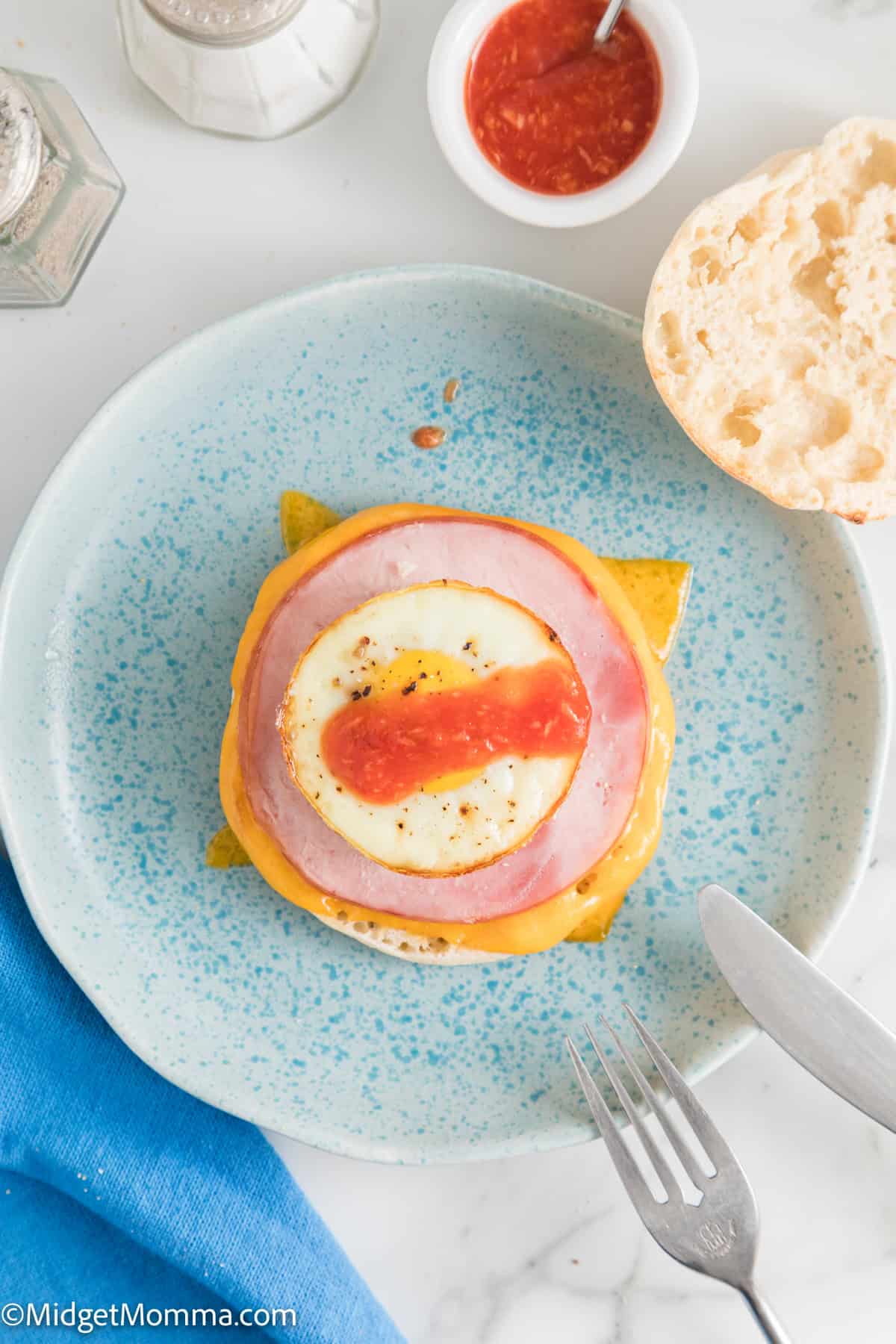 A breakfast sandwich on a blue plate, featuring cheese, ham, egg, and a strip of red sauce. The sandwich's top bun is off to the side. A fork, knife, small bowl of sauce, and salt shaker are also visible.
