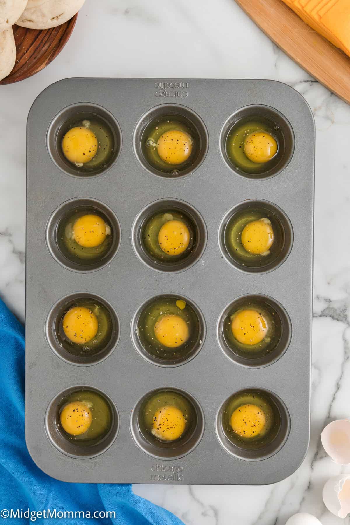 A muffin tin with twelve compartments, each containing a raw egg, is on a marble countertop. A blue cloth and some eggs are visible beside it.