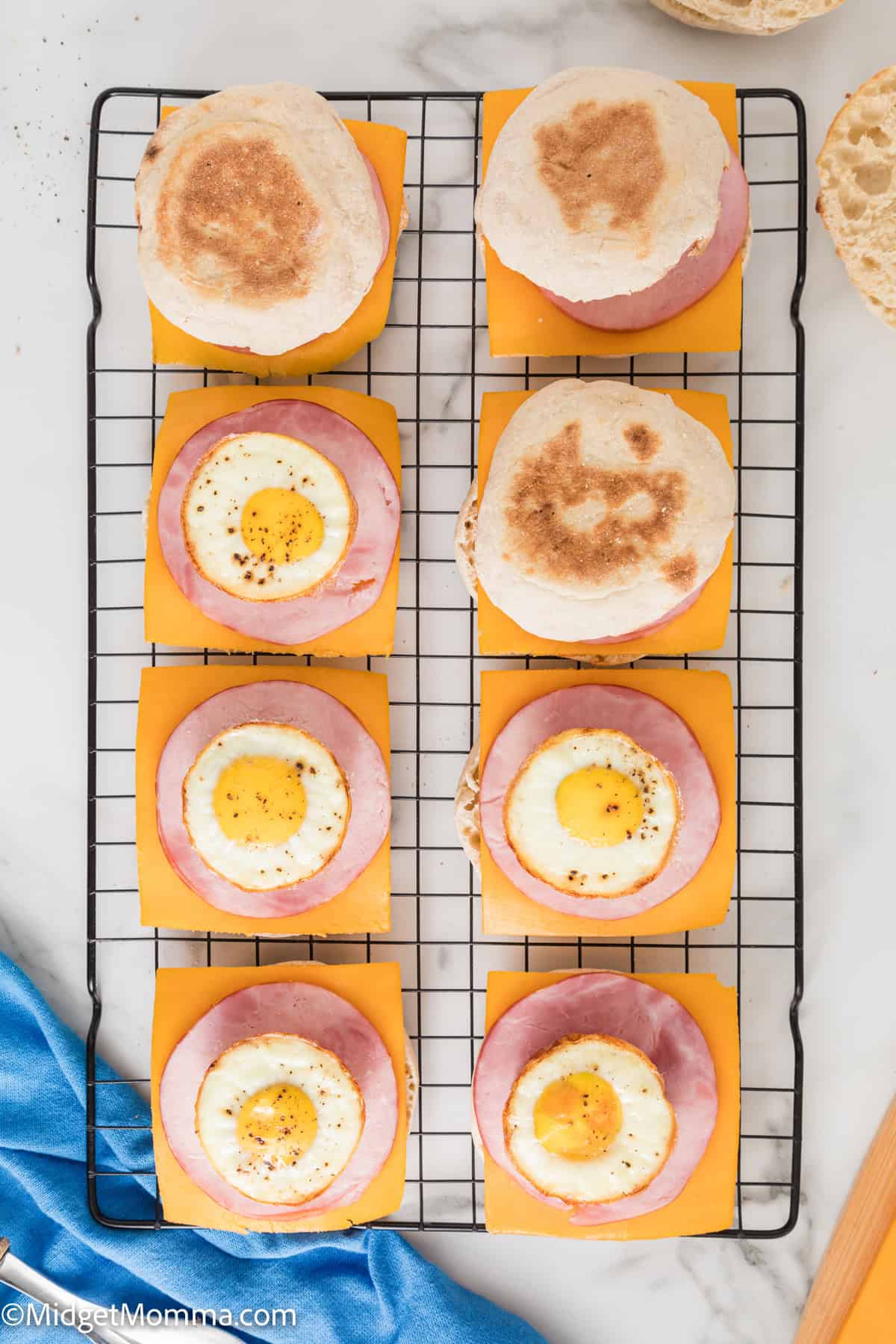 Wire rack holding eight breakfast sandwiches made with English muffins, cheddar cheese, ham, and eggs.