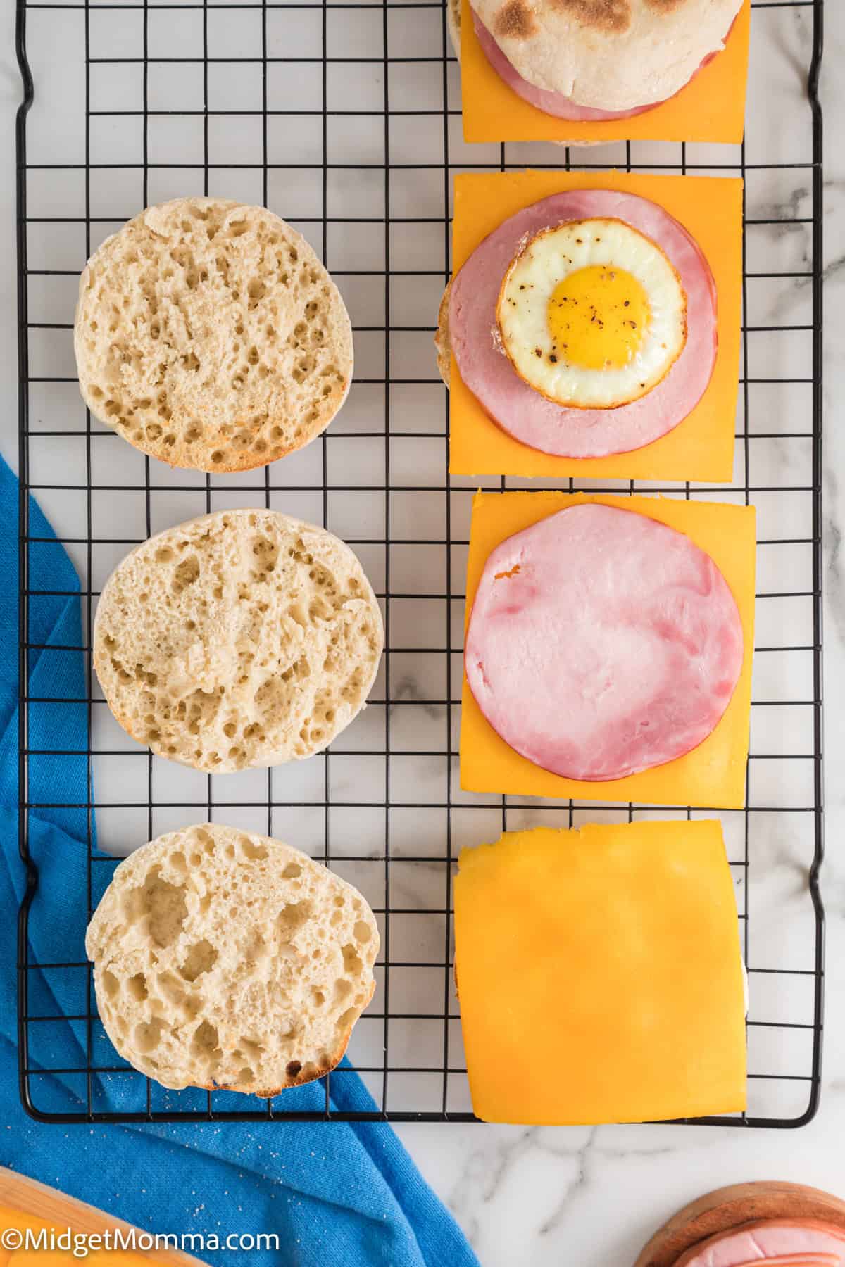 Four open-faced breakfast sandwiches are arranged on a cooling rack. Each sandwich has one half topped with cheese, ham, and a fried egg, and the other half is a plain English muffin.