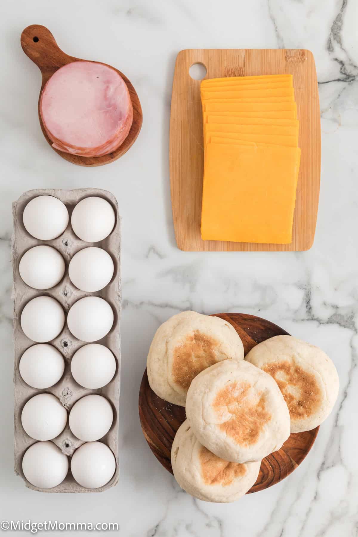 A marble surface with a pack of eggs, a stack of English muffins, a wooden board of cheese slices, and a wooden dish holding a slice of canadian bacon to make egg mcmuffin recipe.