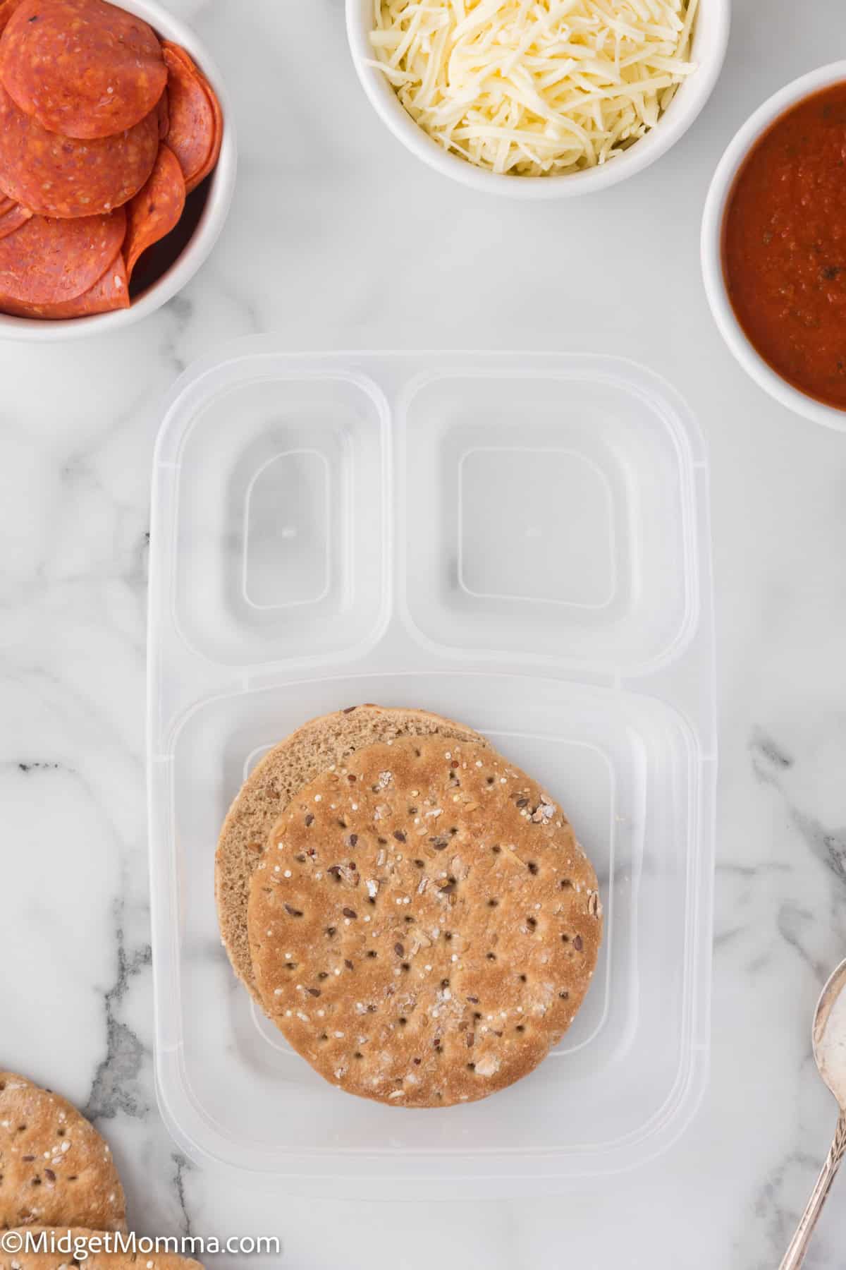 Lunch container with round sandwich thins; side bowls with pepperoni, shredded cheese, and tomato sauce on a marble surface.