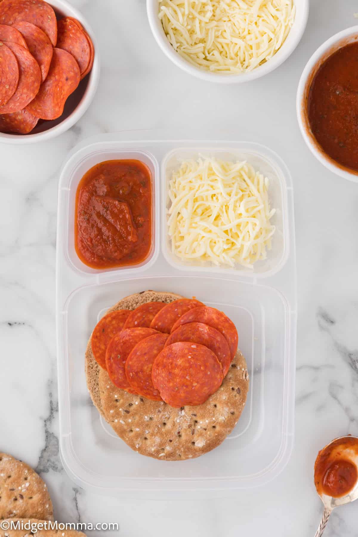 Plastic container with flatbread, pepperoni slices, shredded cheese, and tomato sauce on a marble surface, surrounded by bowls of more ingredients.