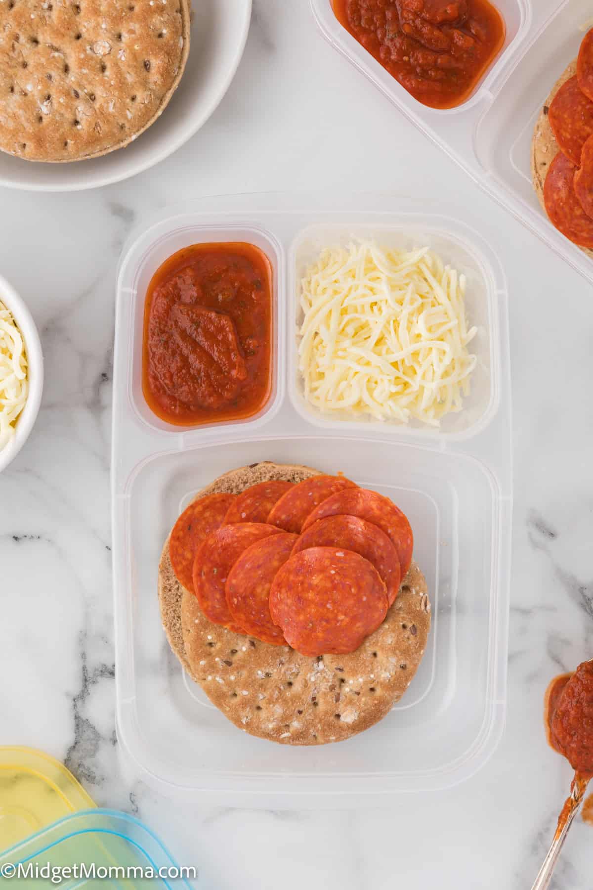 A meal prep container with pizza ingredients: round flatbread, pepperoni slices, shredded cheese, and pizza sauce.