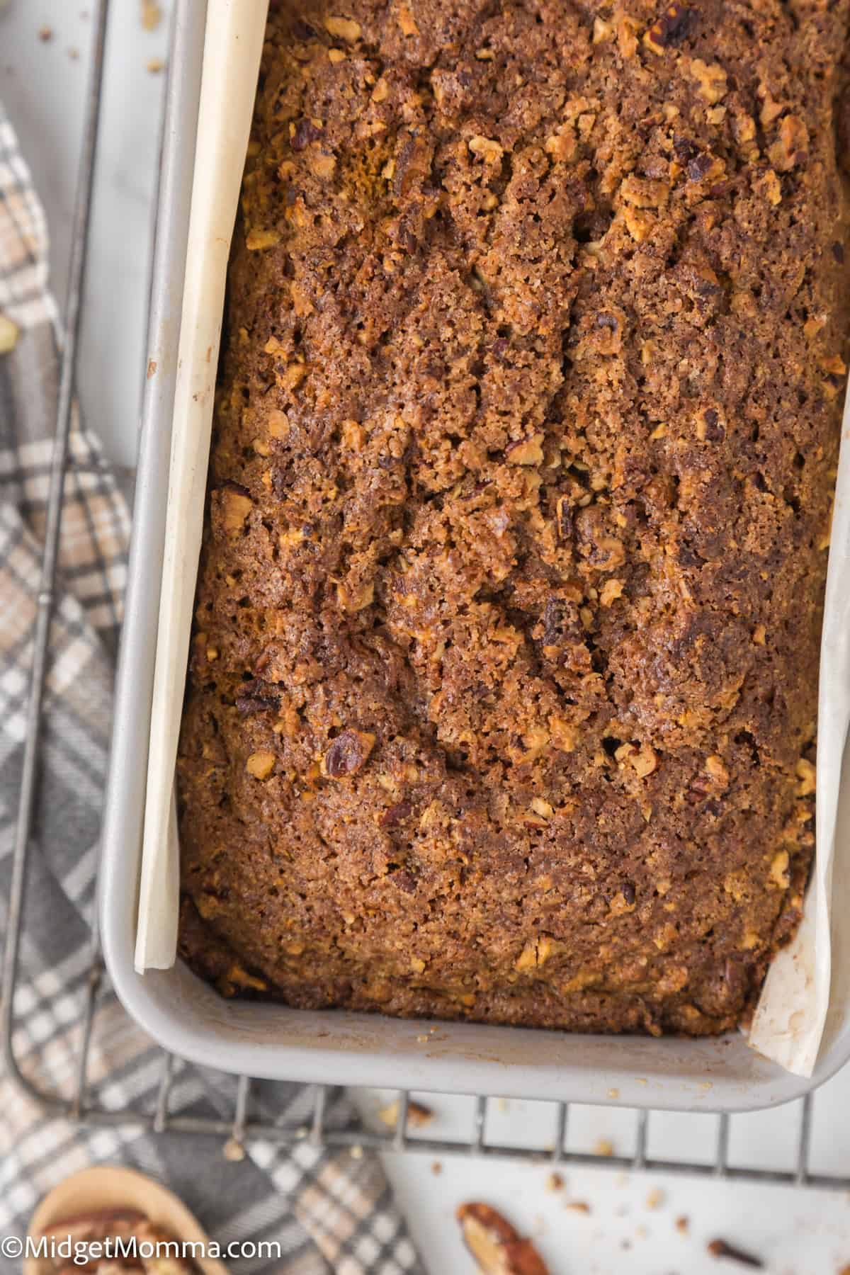 Close-up of a freshly baked loaf of bread in a rectangular tin on a cooling rack. A checkered cloth and scattered crumbs are visible in the background.