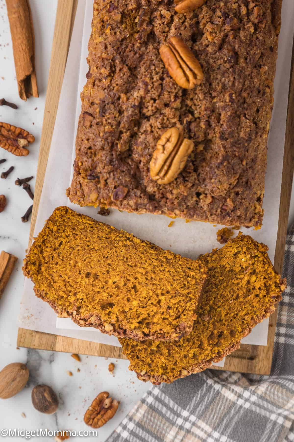 A loaf of pumpkin bread partially sliced, resting on a wooden board. The loaf is topped with pecans, and whole spices are scattered around.