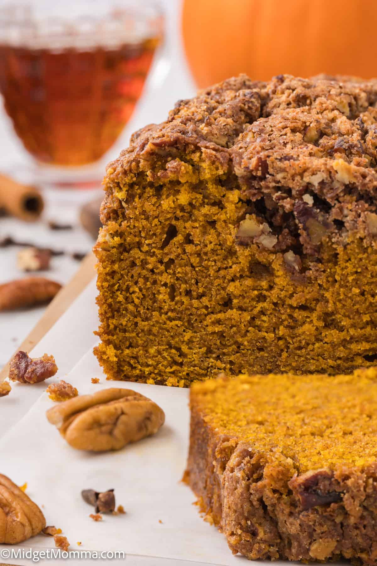 Close-up view of a sliced pumpkin bread with a crumbly topping, pecans, and maple syrup in the background.