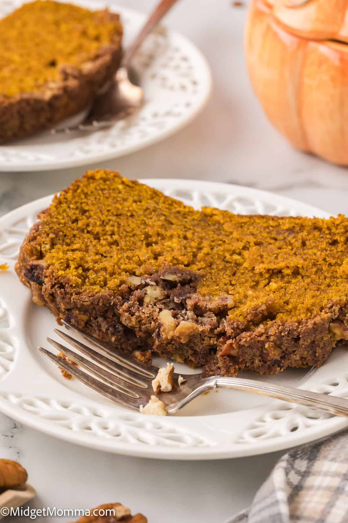 A close-up of two plates of pumpkin pie on a white table. Each plate has a slice of pie with a fork beside it. The pie has a crumbly crust and is garnished with nuts.