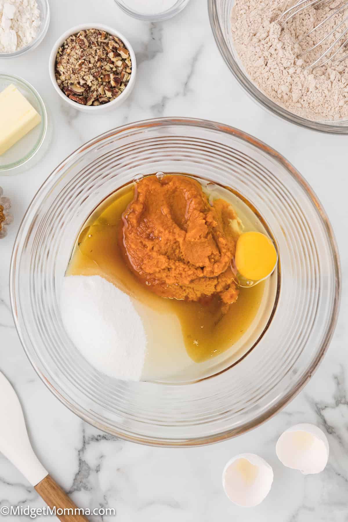 A bowl contains pumpkin puree, sugar, maple syrup, egg and oil ready to be mixed, surrounded by additional baking ingredients on a marble countertop.