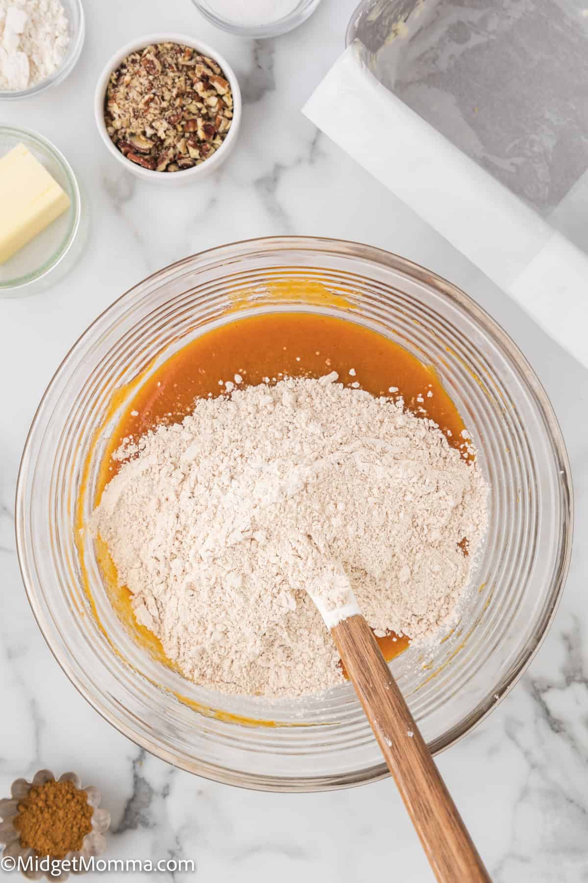 A glass mixing bowl with dry ingredients being stirred into wet ingredients. Nearby are small bowls containing nuts, butter, and spices. A lined baking pan is partially visible in the corner.