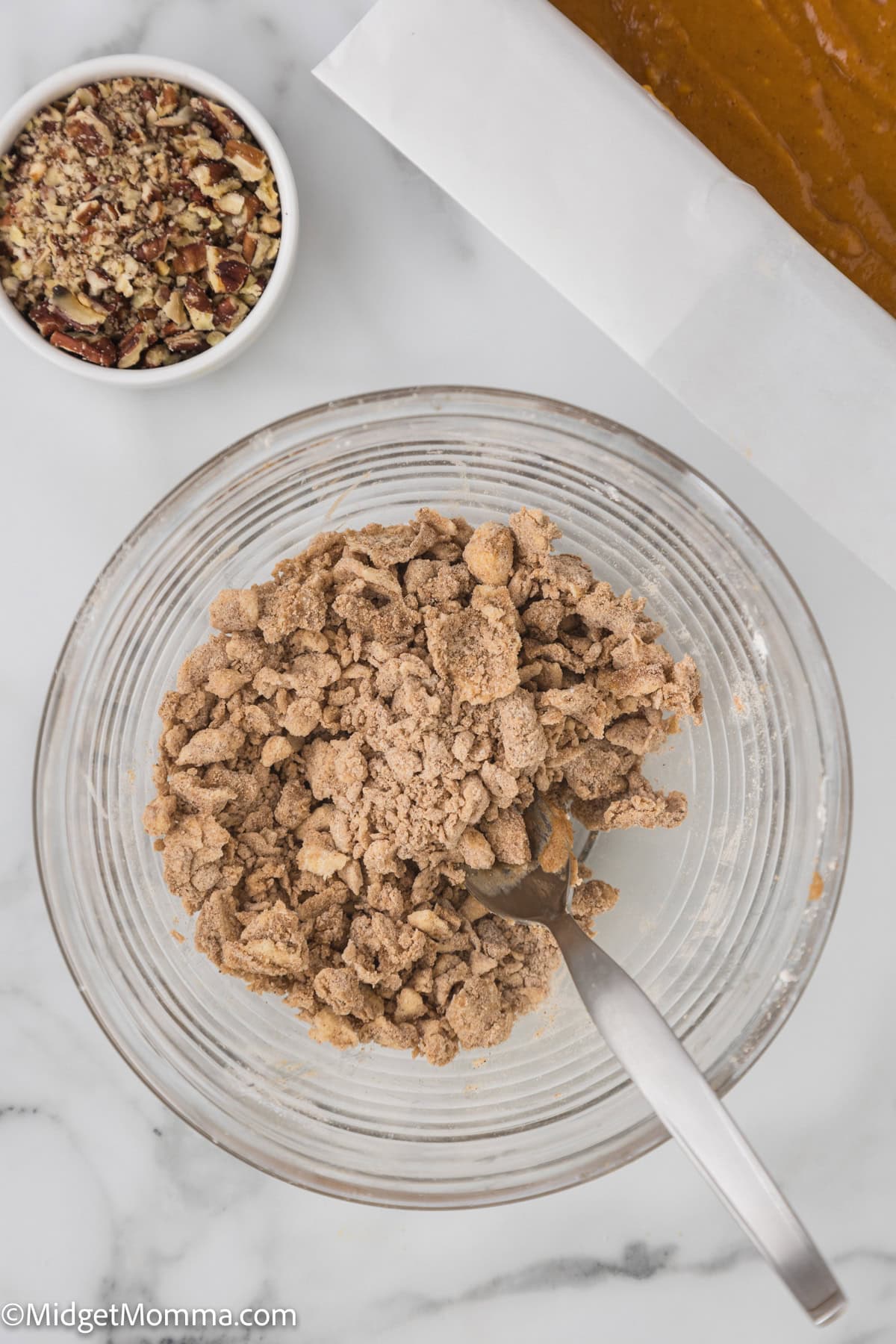 A clear glass bowl crumbly, struesel topping mixture and a silver spoon, placed on a marble countertop. A small white bowl with chopped nuts is positioned nearby, next to a parchment-lined baking dish.