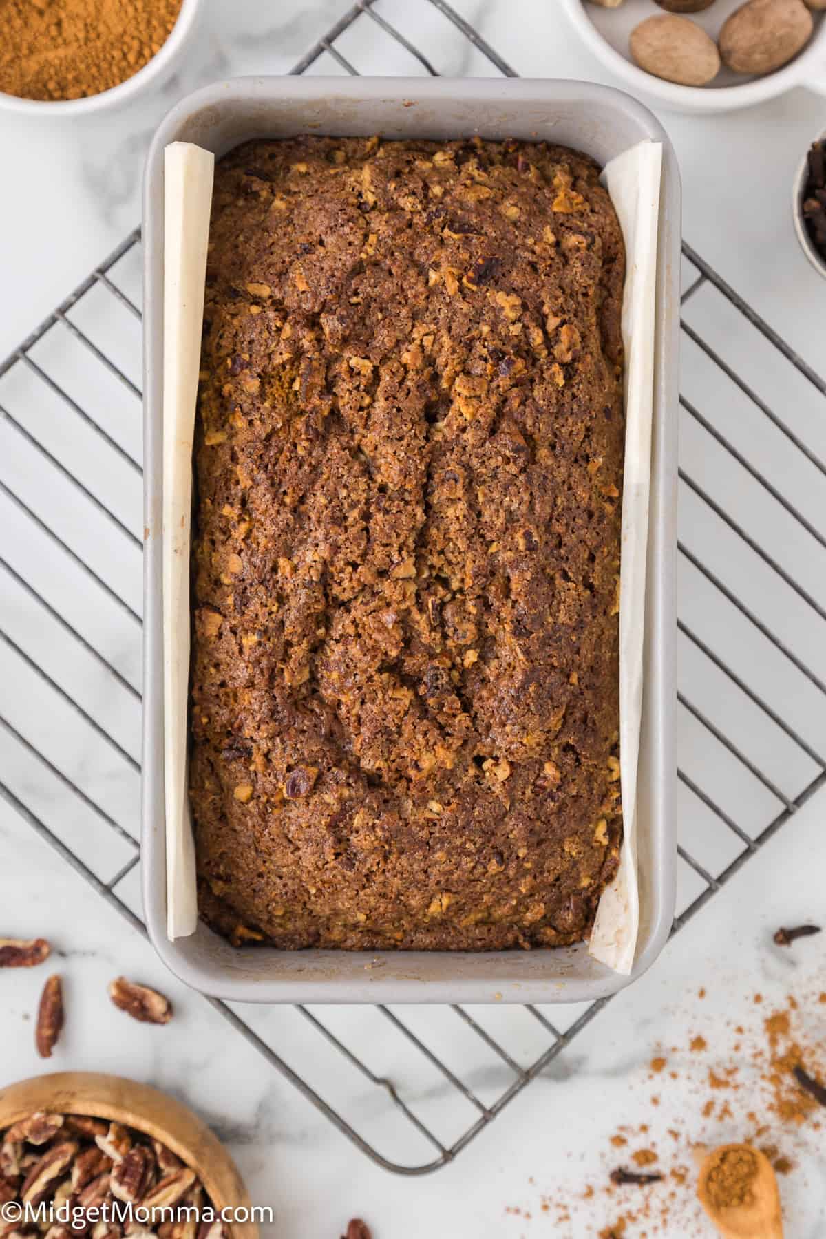 A freshly baked loaf of banana bread in a rectangular pan placed on a wire cooling rack, with assorted ingredients like pecans, nutmeg, and cinnamon surrounding it.