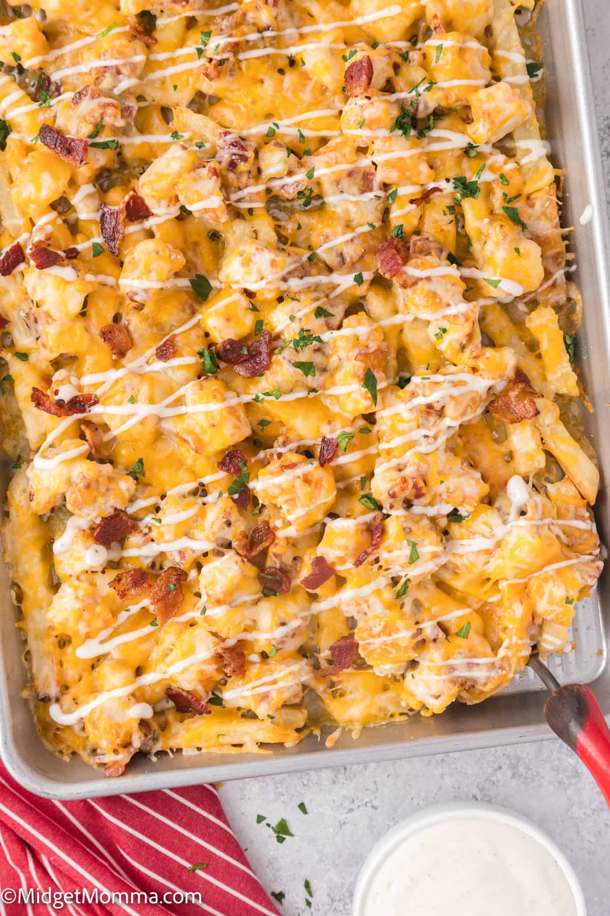 A baking tray filled with loaded cheese and bacon french fries, topped with a drizzle of ranch sauce and garnished with chopped herbs. A red cloth is on the side.