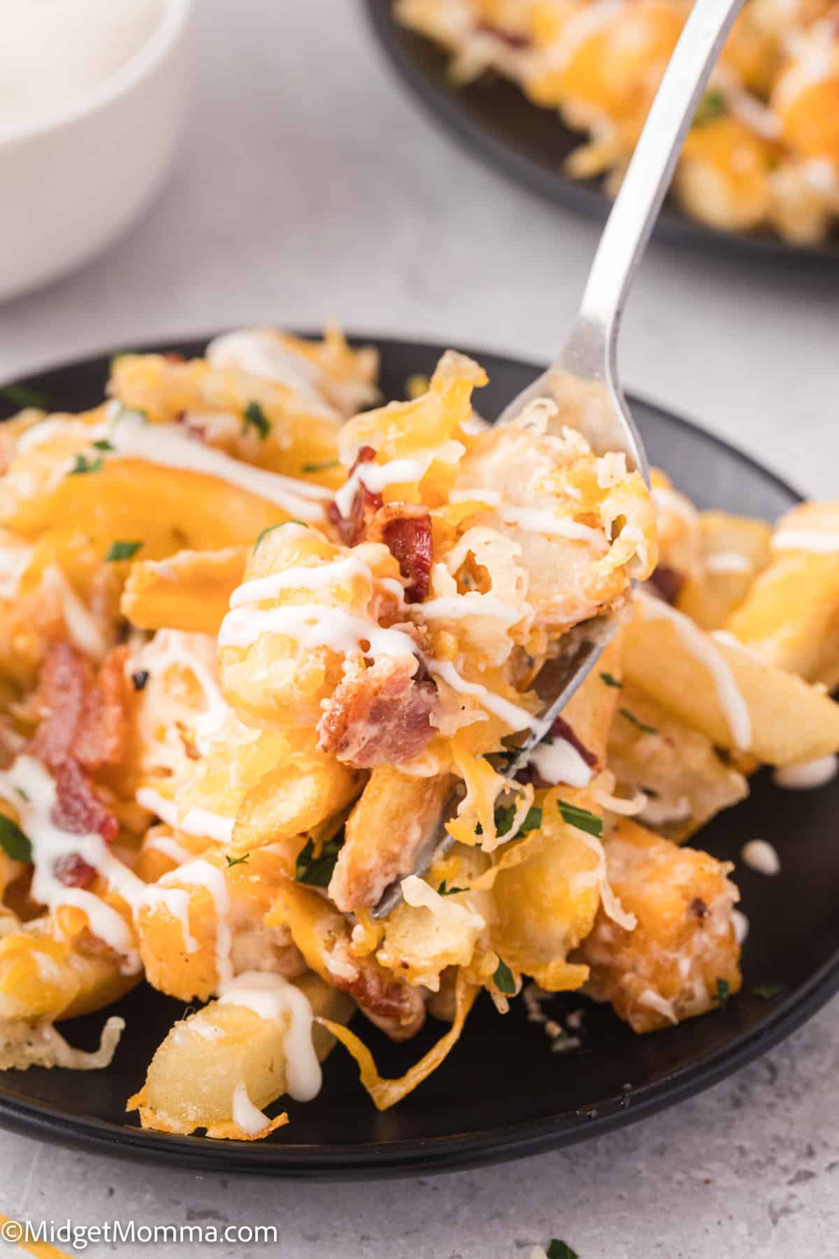 Close-up of a fork holding cheesy loaded fries topped with bacon bits and sauce on a black plate.