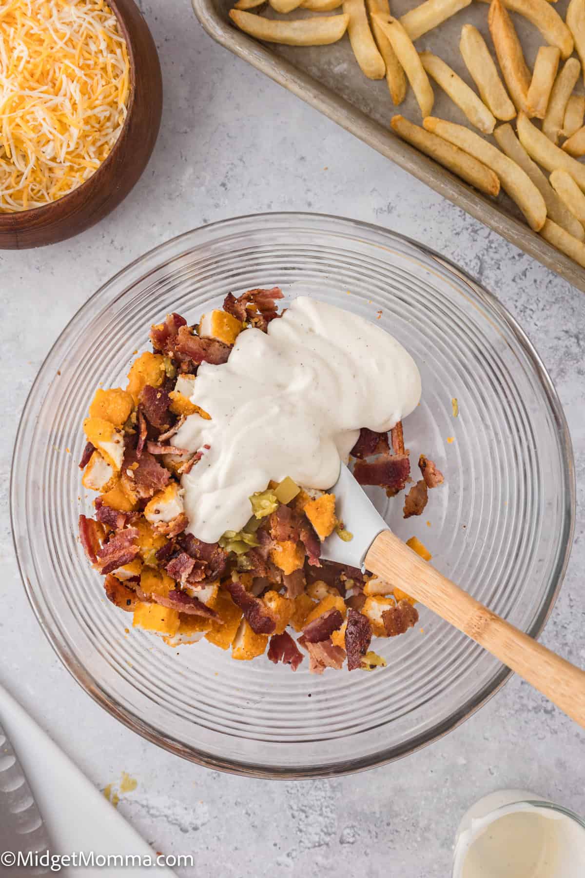 A clear bowl with diced bacon, chopped chicken, and ranch dressing sauce. A wooden spatula rests in the mixture. Adjacent, fries on a baking sheet and shredded cheese in a bowl.