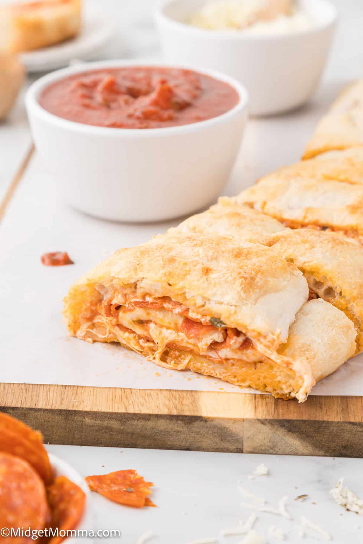 A sliced pizza bread on a wooden board, with visible layers of meat and cheese, is in the foreground. In the background, there is a small bowl of marinara sauce.