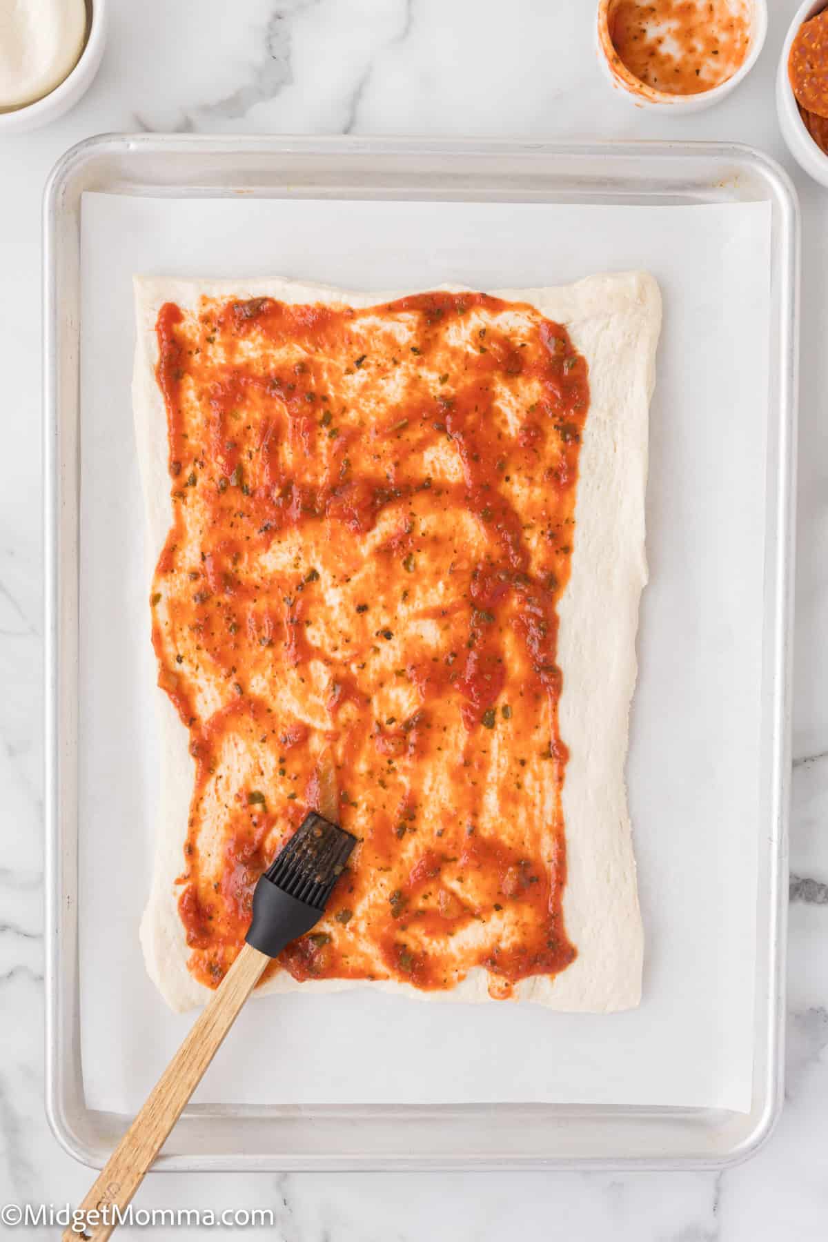 A rectangular sheet of dough on a baking tray is spread with tomato sauce using a brush.