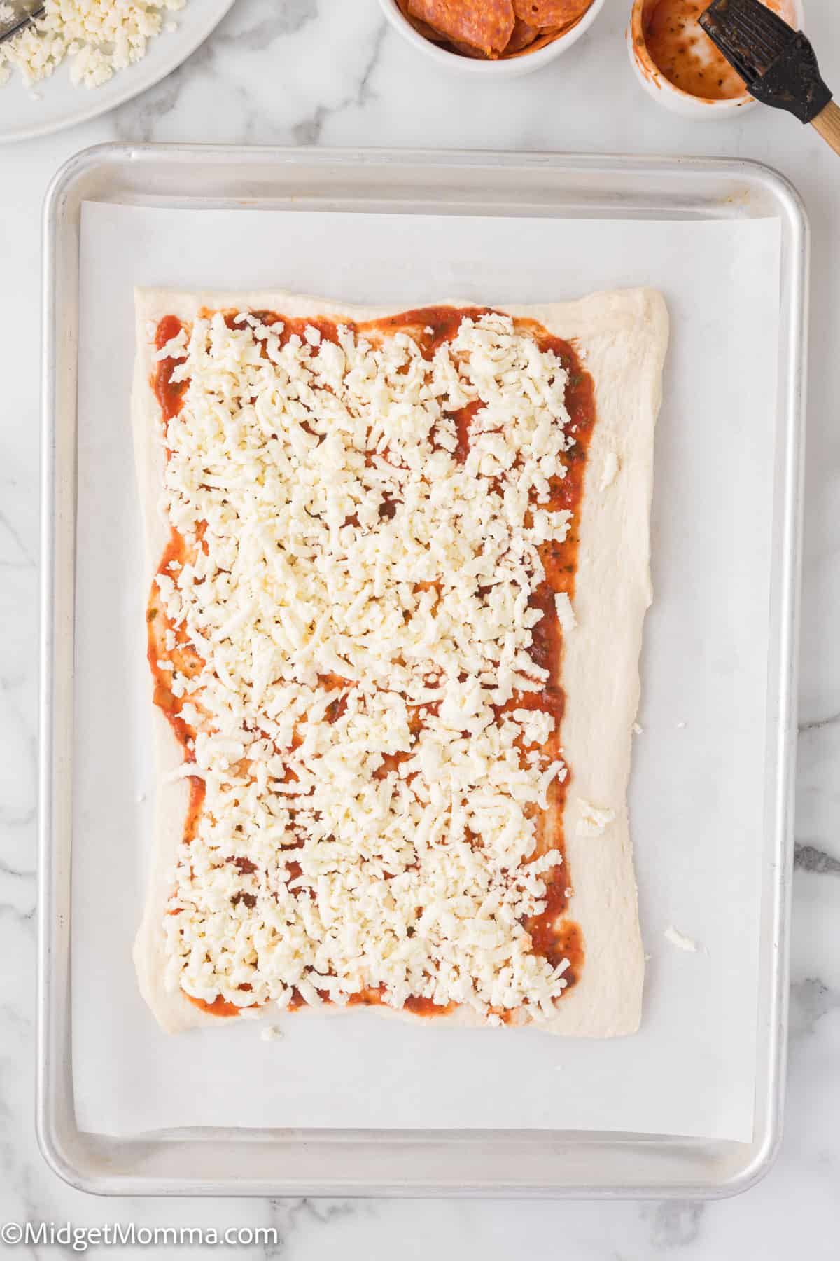 Rectangle pizza dough with tomato sauce and shredded cheese on a baking sheet. Small bowls with ingredients in the background.