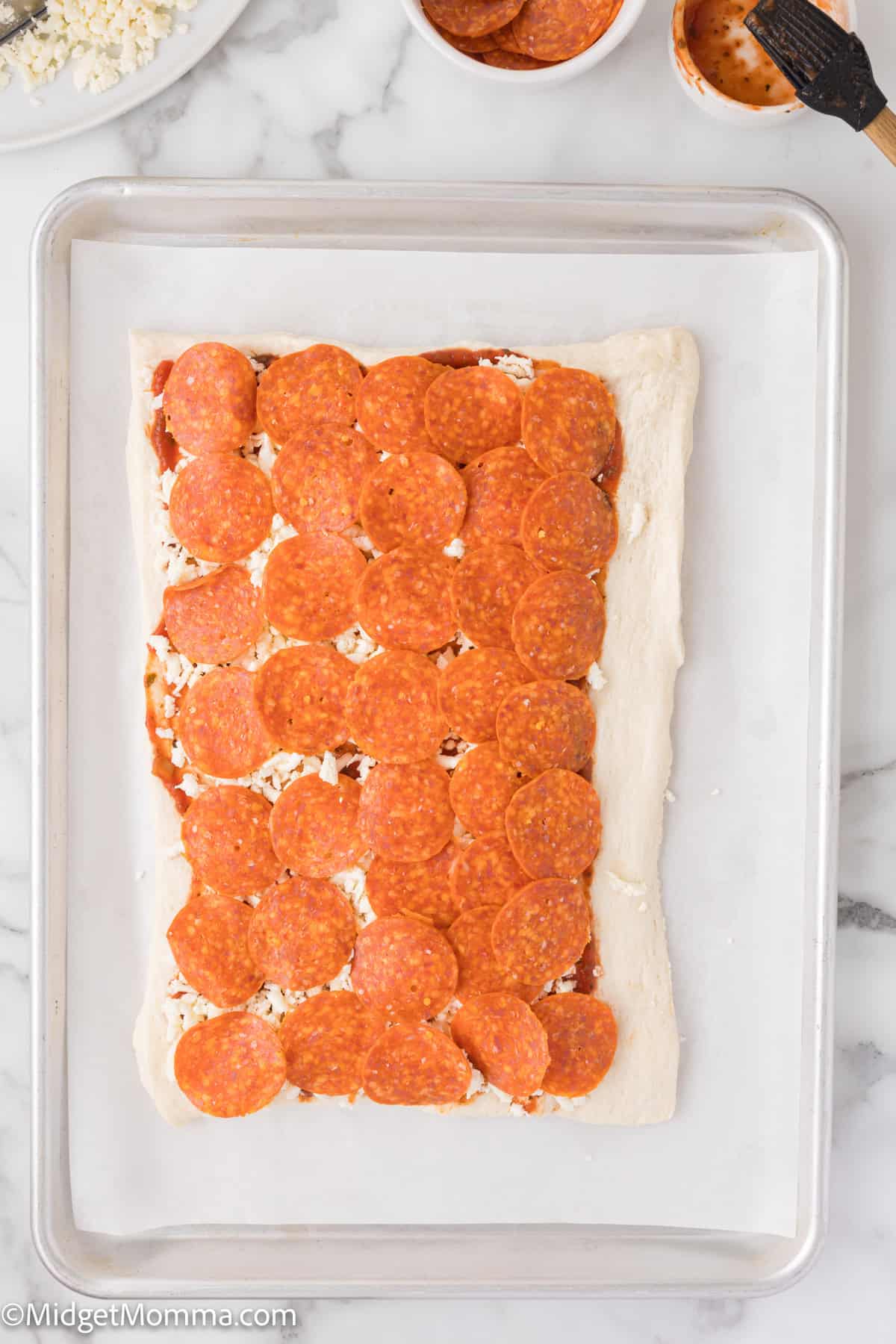 Rectangular dough topped with tomato sauce, cheese, and pepperoni slices on a baking tray.