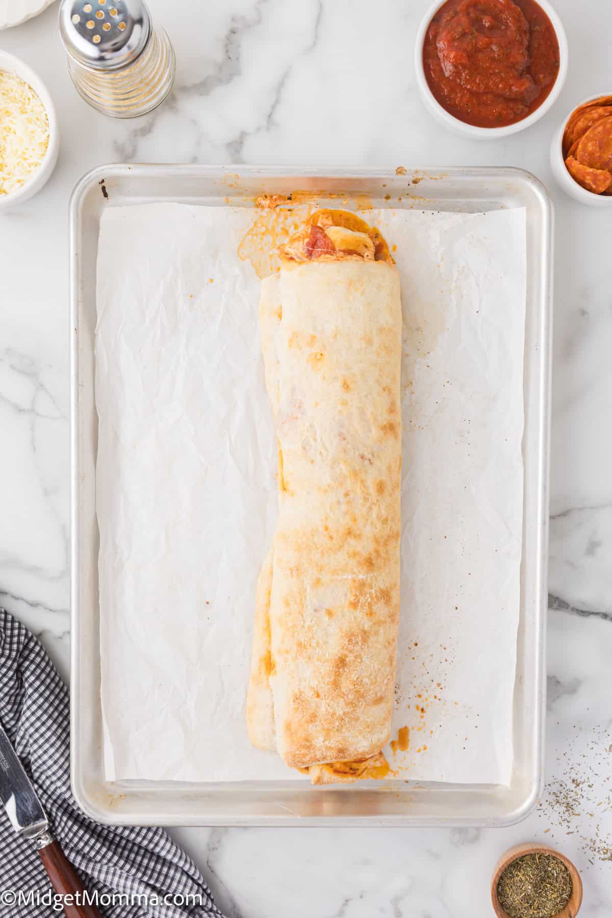 A rolled dough with a golden brown top on a baking sheet, surrounded by small bowls of ingredients and a pepper grinder.