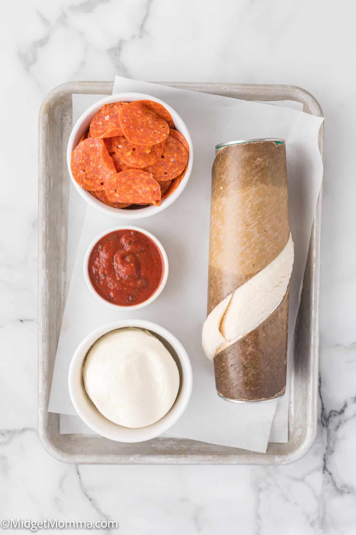 Tray with ingredients: a can of pizza dough, a bowl of pepperoni slices, a bowl of tomato sauce, and a bowl of mozzarella cheese on parchment paper.