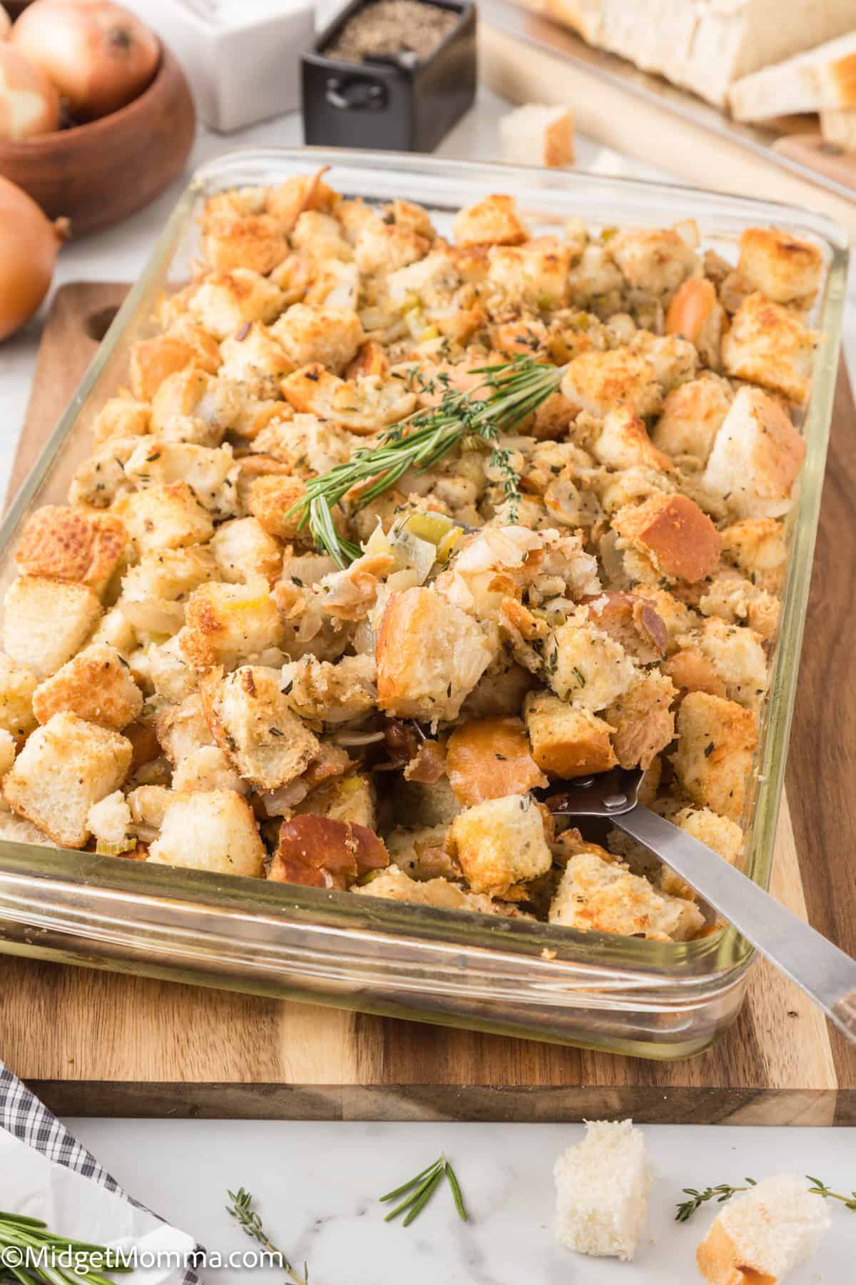 A glass dish filled with bread stuffing, garnished with herbs, sits on a wooden board. Nearby are onions, spices, and sliced bread.