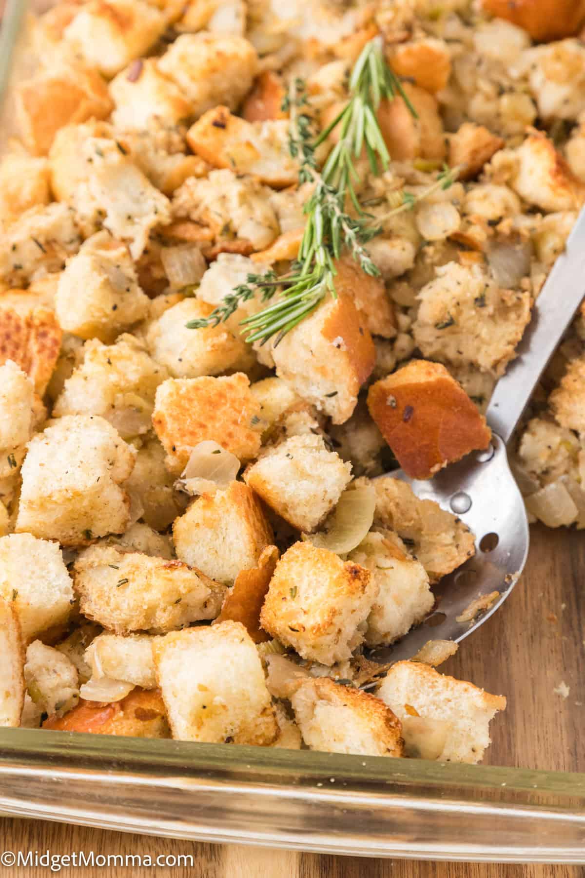 A tray of seasoned bread stuffing with a serving spoon, garnished with sprigs of fresh herbs.