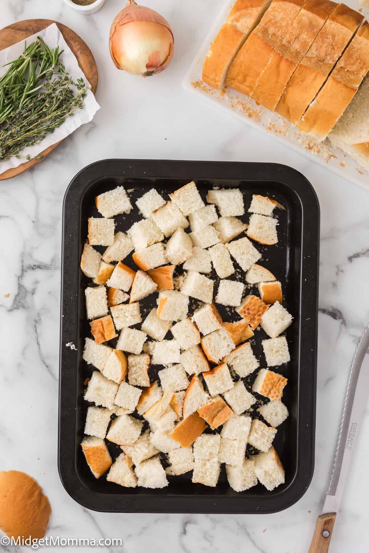 Tray of cubed bread pieces on a marble countertop, surrounded by ingredients like herbs, an onion, and a loaf of sliced bread.