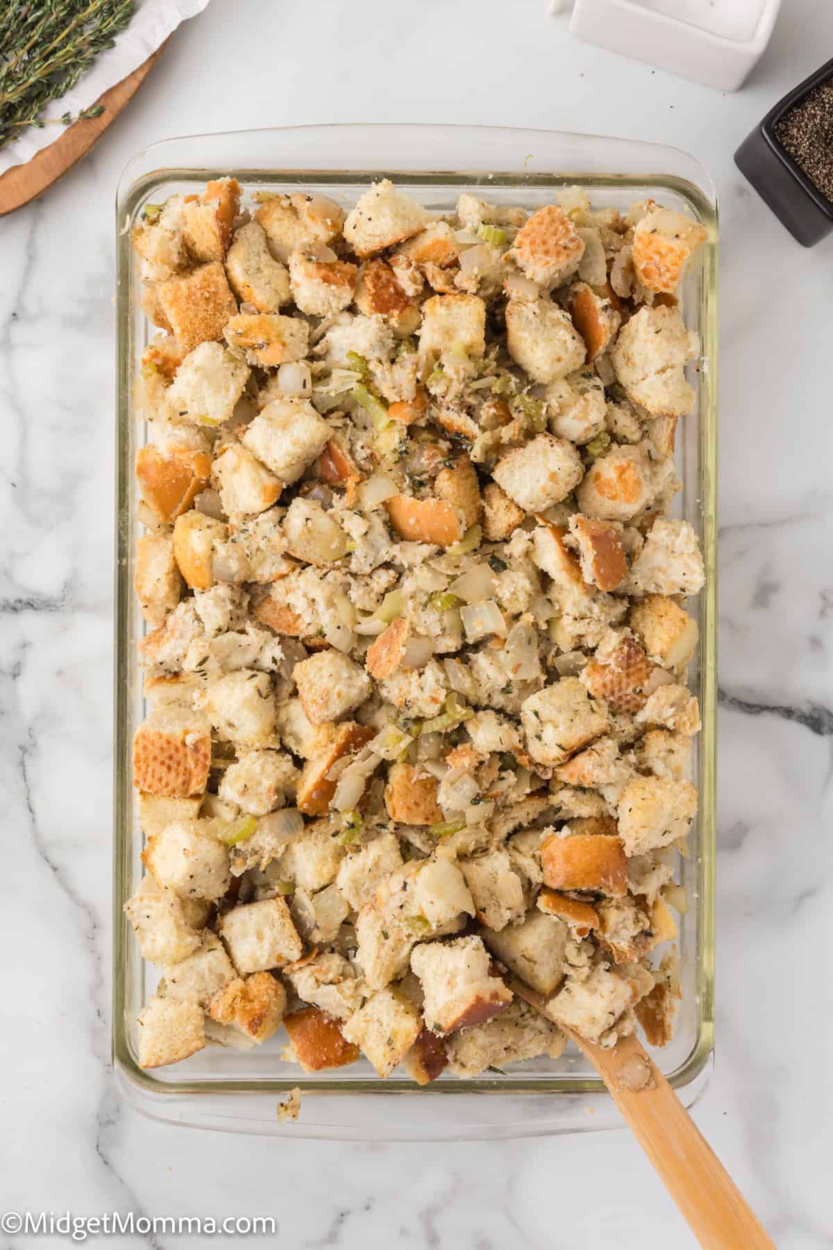 A glass baking dish filled with cubed bread stuffing mixed with celery and herbs, on a marble surface.