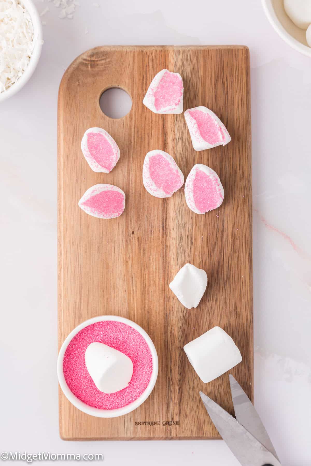 Wooden board with marshmallows cut in half, some dipped in pink sugar. Bowl of pink sugar and scissors nearby.