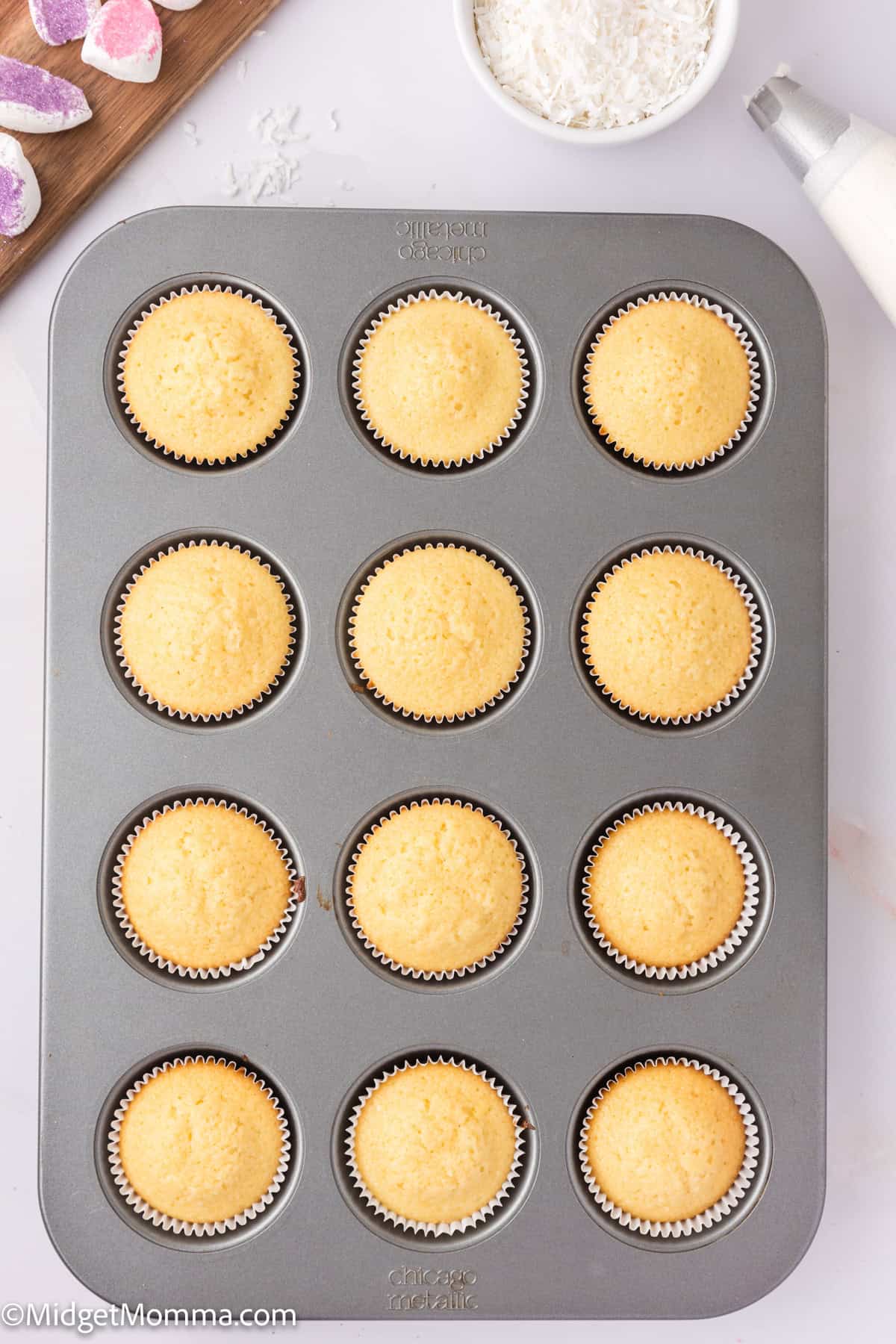 A baking tray with twelve plain cupcakes in paper liners, situated on a white surface. Nearby, there's a piping bag and a bowl of white icing.