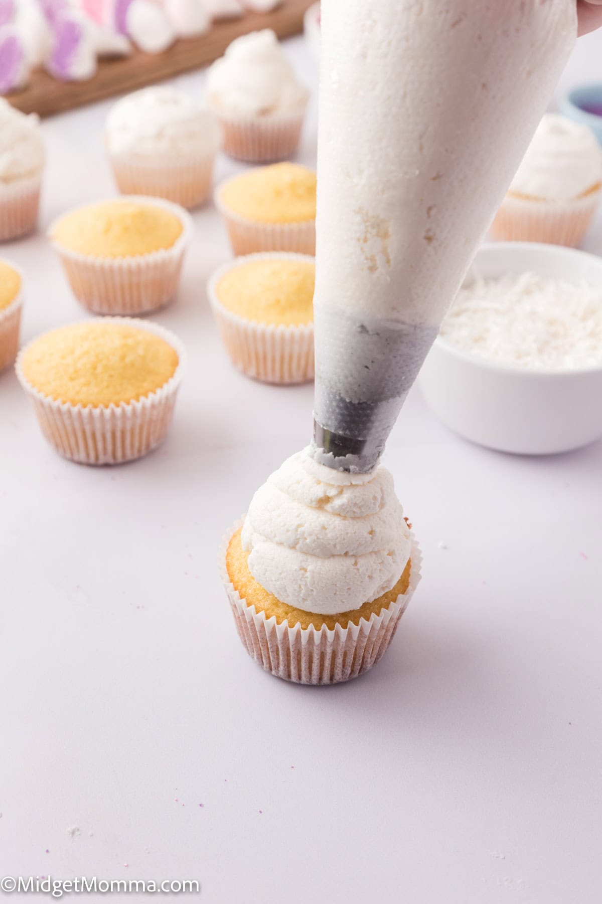 Person piping white frosting onto a vanilla cupcake, surrounded by unfrosted cupcakes and a bowl of frosting in the background.