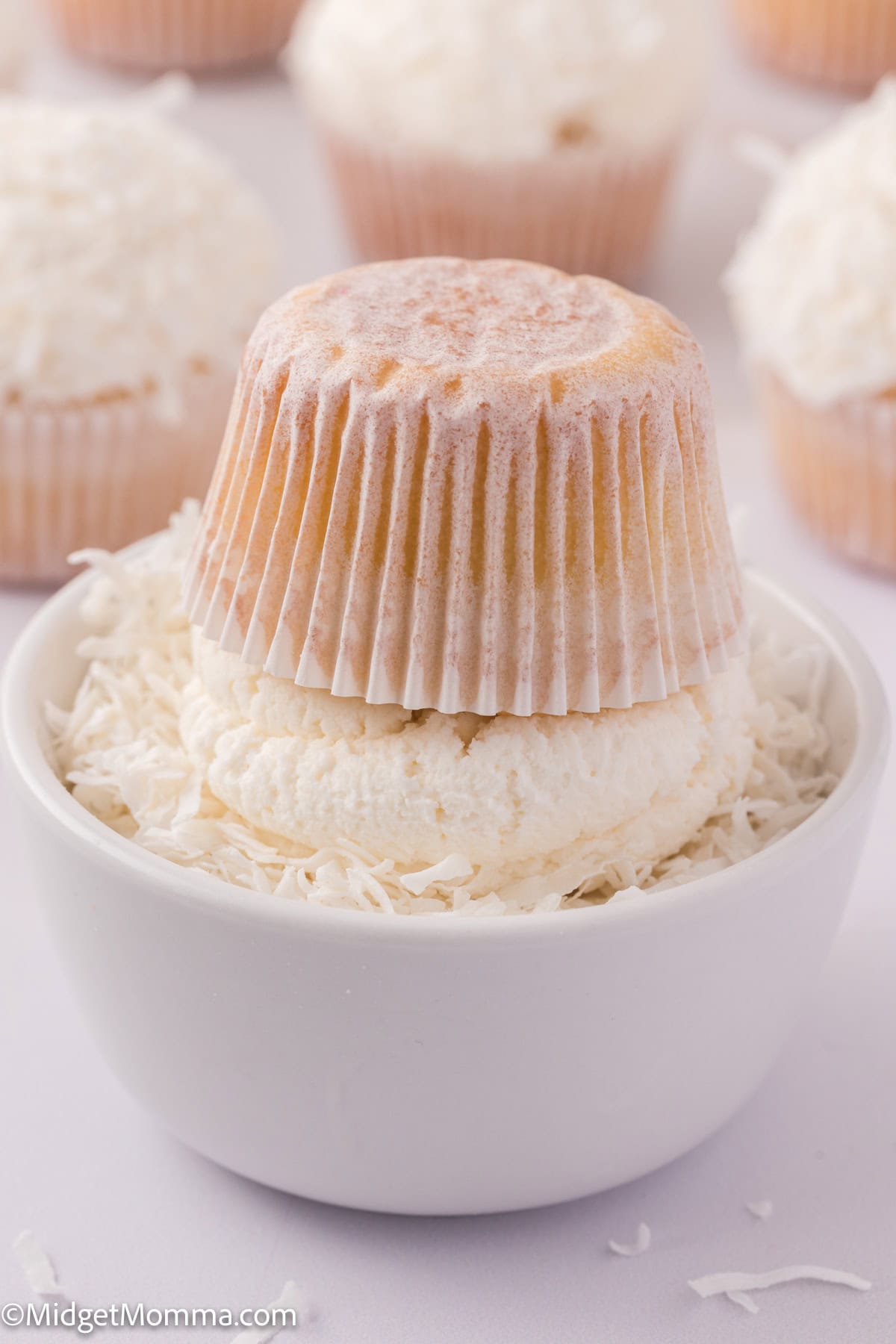 Cupcake upside down in a bowl with shaved coconut, surrounded by more cupcakes topped with coconut shavings.