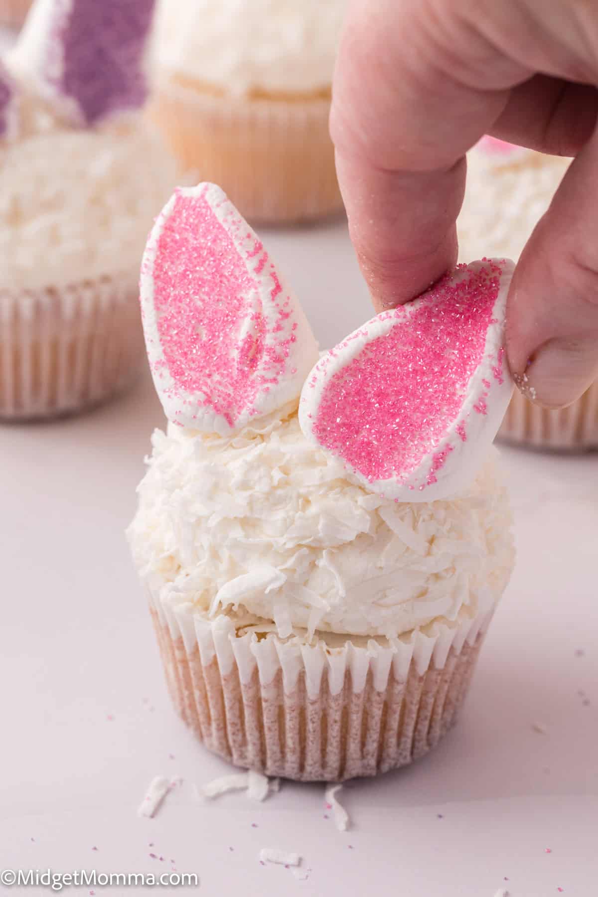 A hand placing pink sugar-coated marshmallow ears on a white frosted cupcake, resembling a bunny.