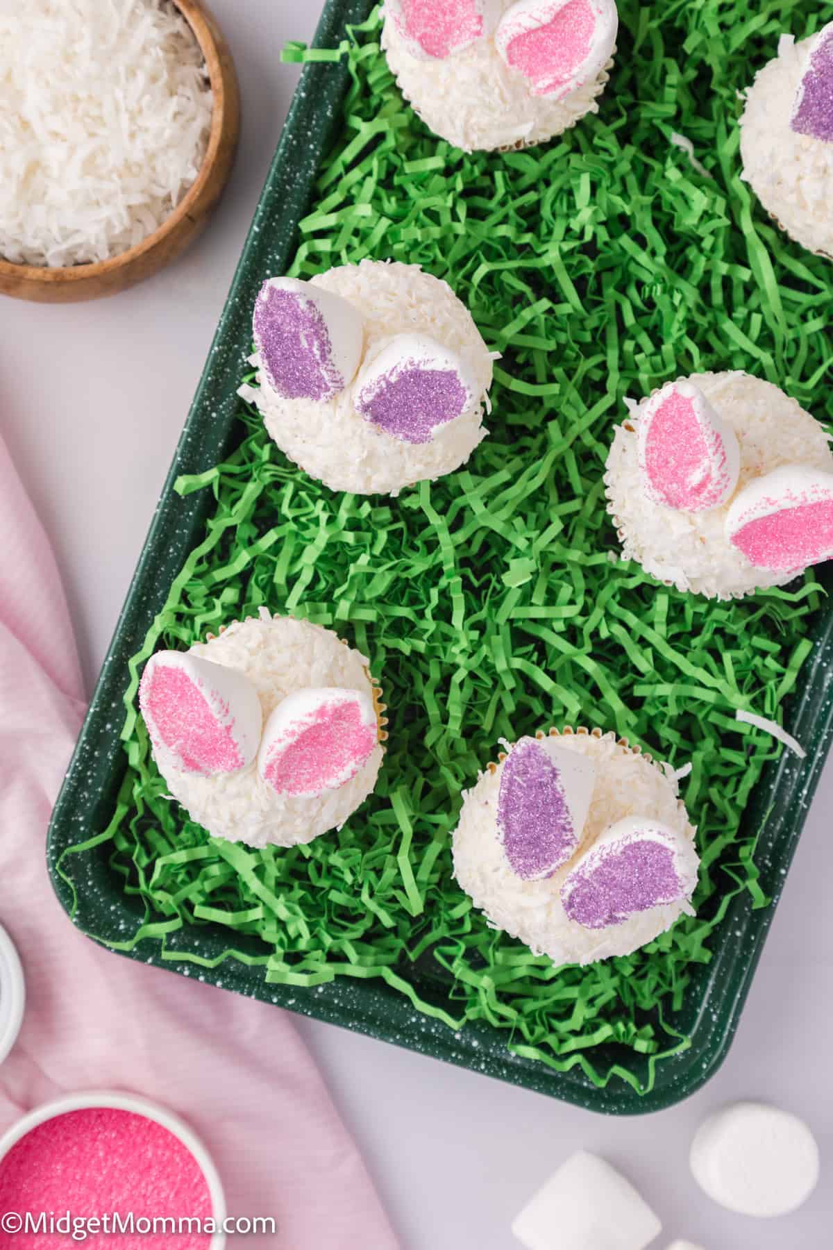 Cupcakes decorated with coconut flakes and pastel pink and purple bunny ears made of candy are displayed on a tray with green shredded paper, resembling grass.