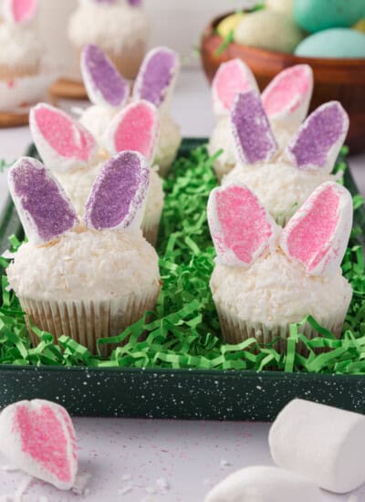 Cupcakes with bunny ear decorations in pink and purple, topped with white frosting and surrounded by green grass-like decor.