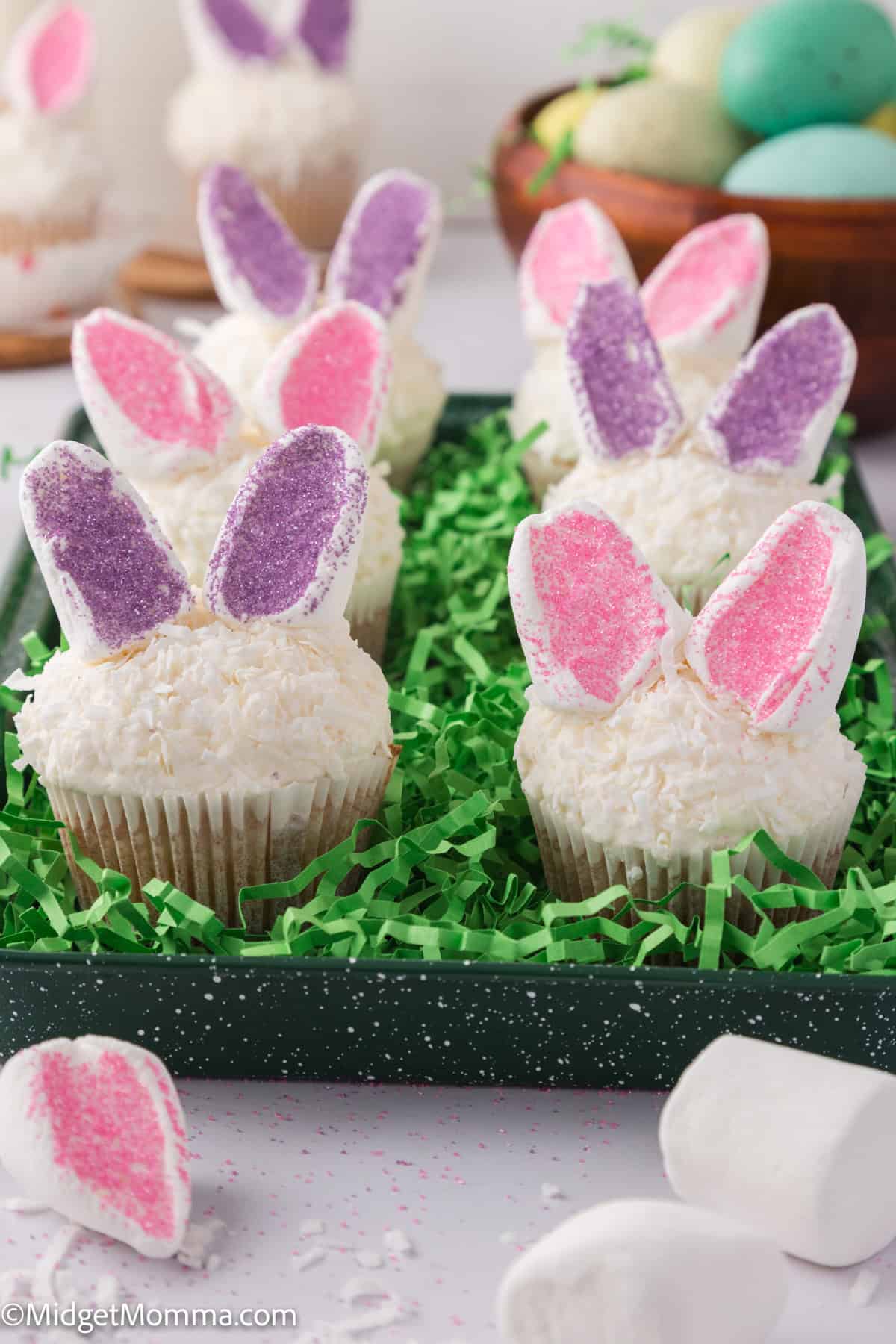 Cupcakes with bunny ear decorations in pink and purple, topped with white frosting and surrounded by green grass-like decor.