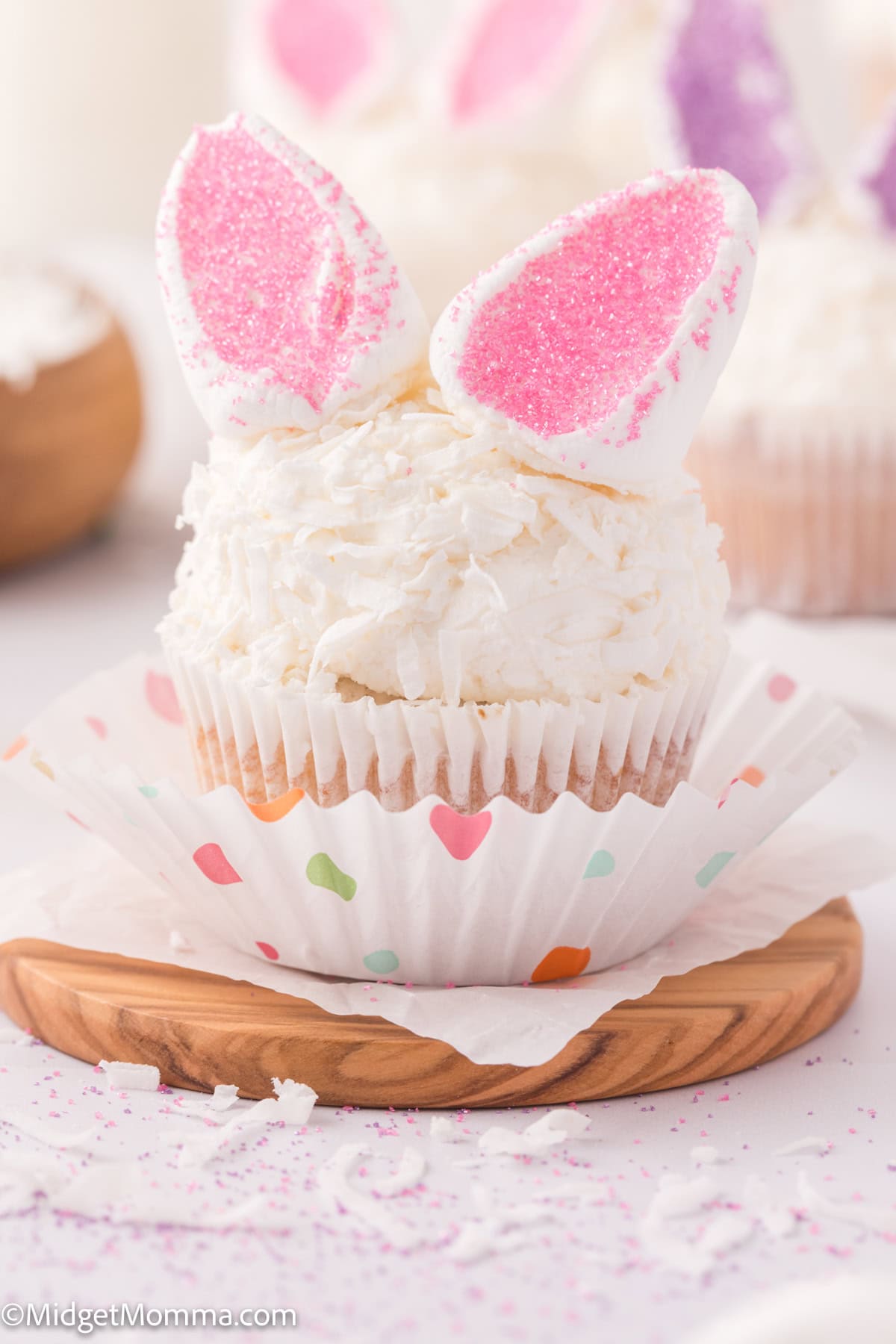 A cupcake with white frosting, topped with marshmallow halves shaped like bunny ears sprinkled with pink sugar, sits on a wooden plate. It's wrapped in a heart-patterned paper liner.