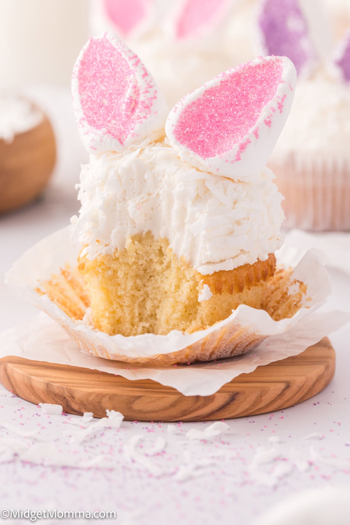 A cupcake with white frosting and pink bunny ear decorations, partially eaten, sits on a wooden plate with a crumpled wrapper.