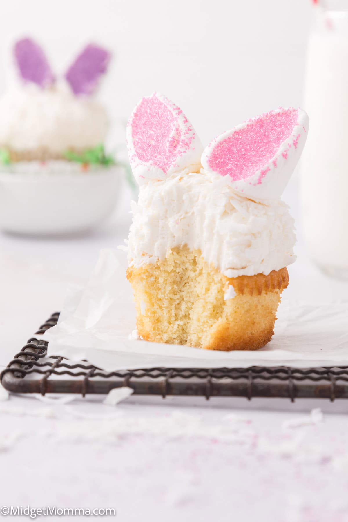 Cupcake topped with white frosting and pink sugar-coated marshmallow bunny ears, with a bite taken out.