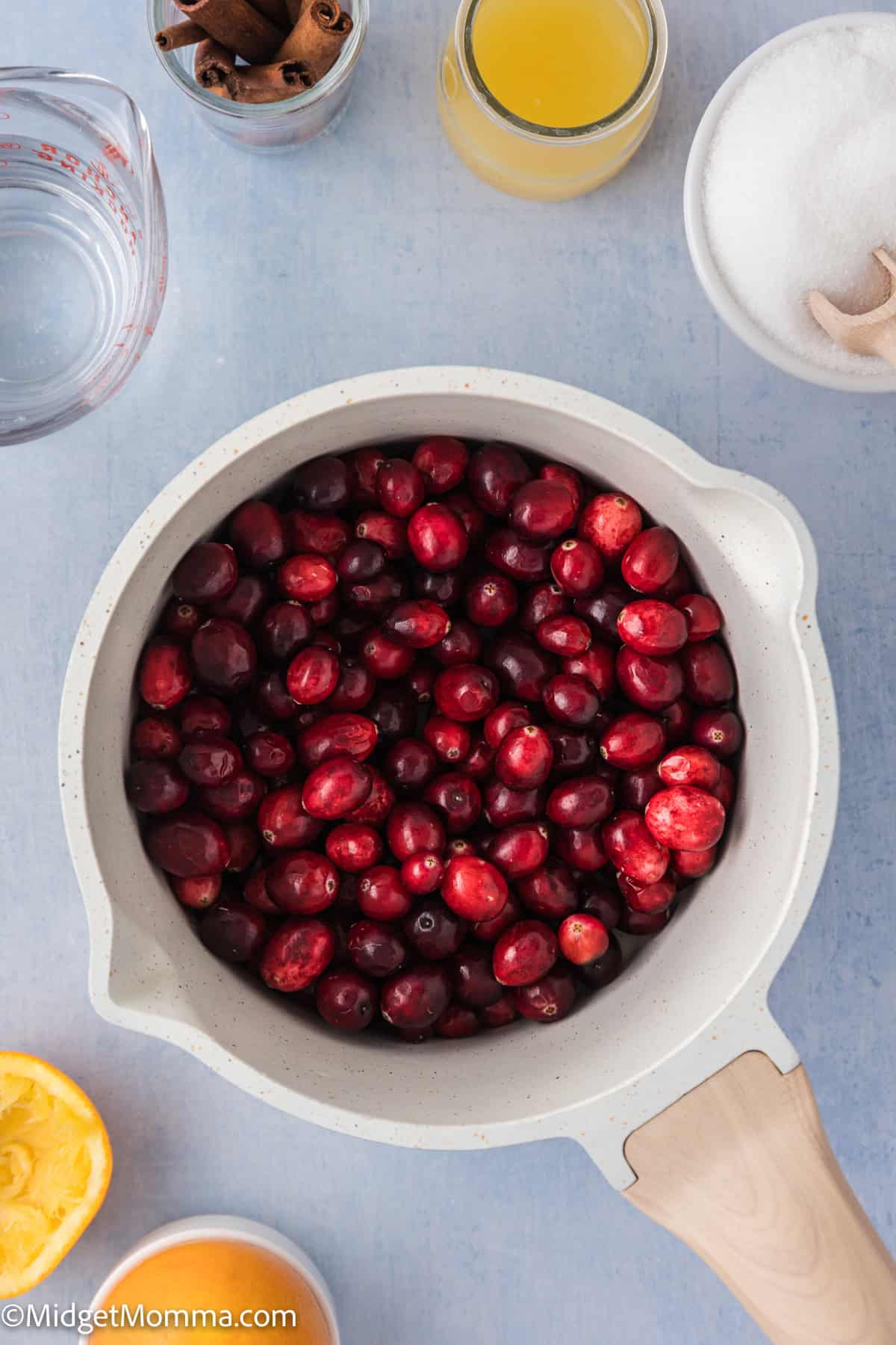 A pot filled with fresh cranberries is surrounded by glasses of water and orange juice, a bowl of sugar, cinnamon sticks, and half an orange on a light blue surface.