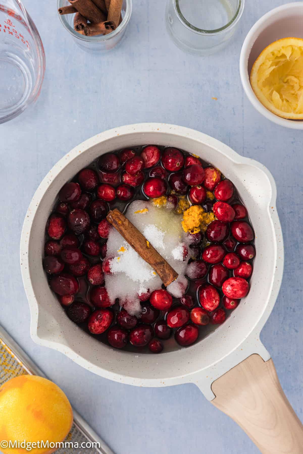 Pot filled with cranberries, sugar, orange zest, and cinnamon stick on a light blue surface. Nearby are ingredients in bowls and jars.