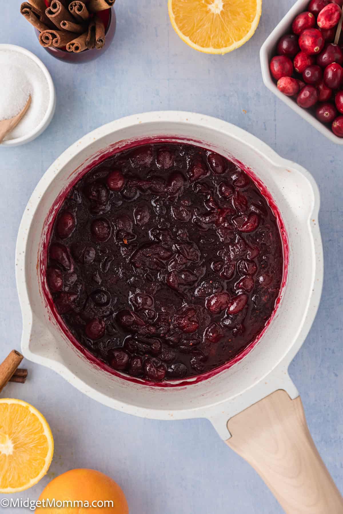 A pot of cooked cranberry sauce is surrounded by fresh cranberries, cinnamon sticks, a halved orange, and a bowl of sugar on a light blue surface.