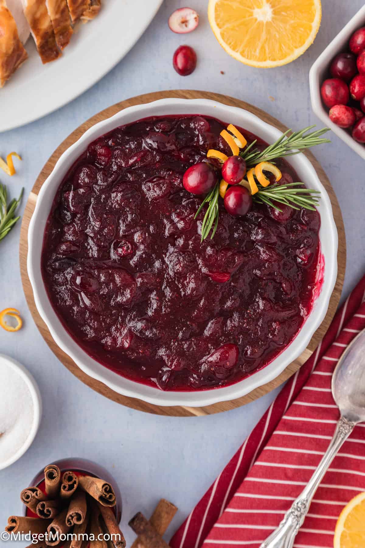 A bowl of cranberry sauce garnished with rosemary and orange peel. Nearby are orange slices, whole cranberries, cinnamon sticks, and a red-striped napkin with a spoon.