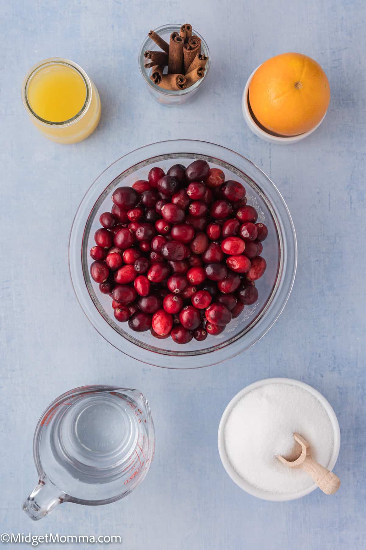 A bowl of cranberries surrounded by a glass of juice, cinnamon sticks in a jar, an orange, a bowl of sugar with a spoon, and a pitcher of water on a light blue surface.