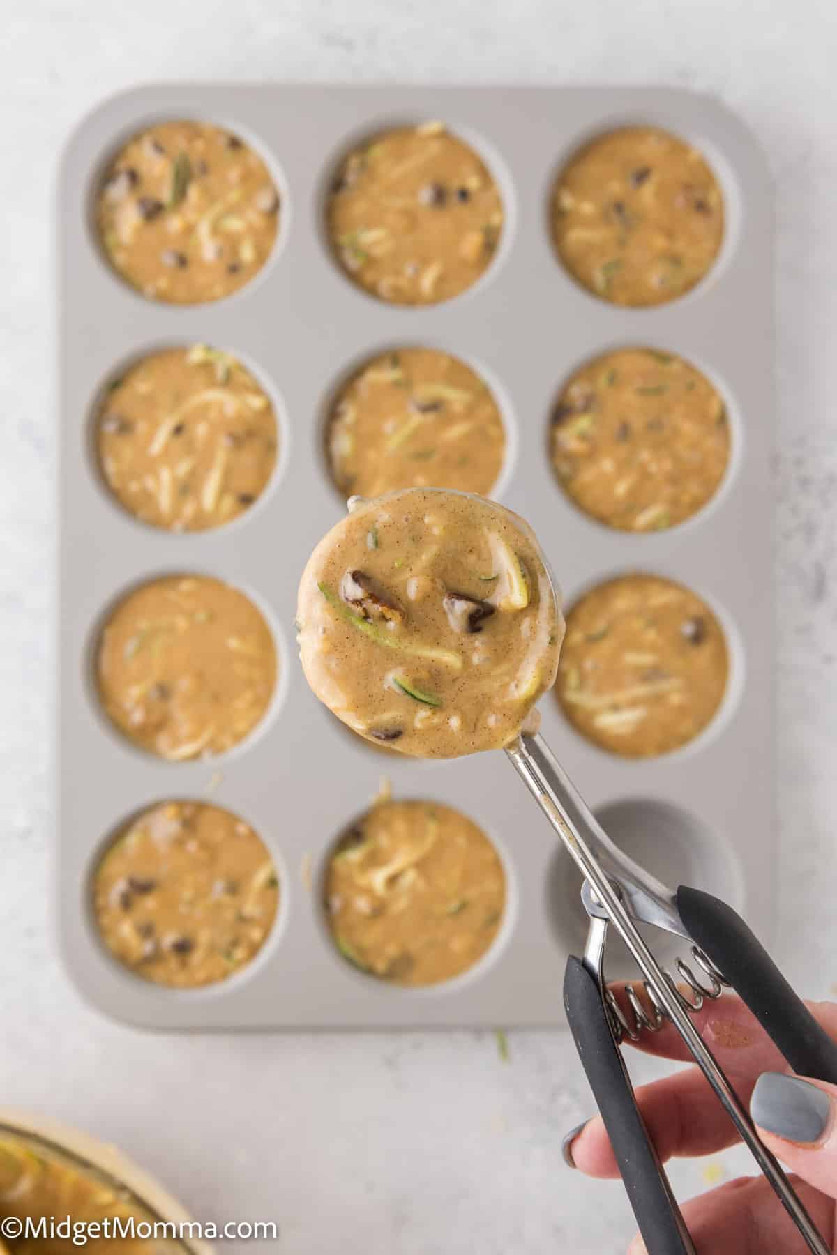 Muffin batter with chocolate chips and zucchini being scooped into a muffin tin using a cookie scoop.