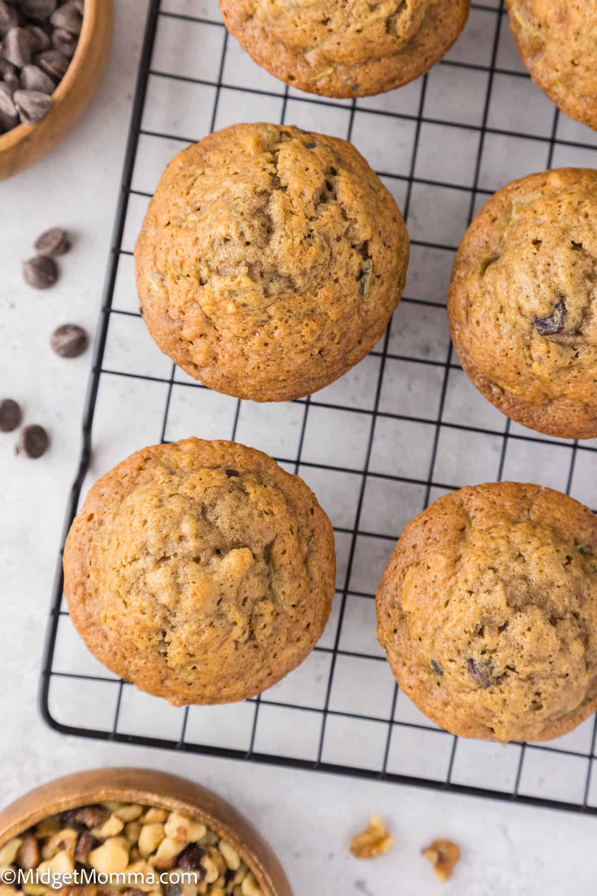 Six muffins cooling on a black wire rack, surrounded by bowls of chocolate chips and nuts on a light surface.