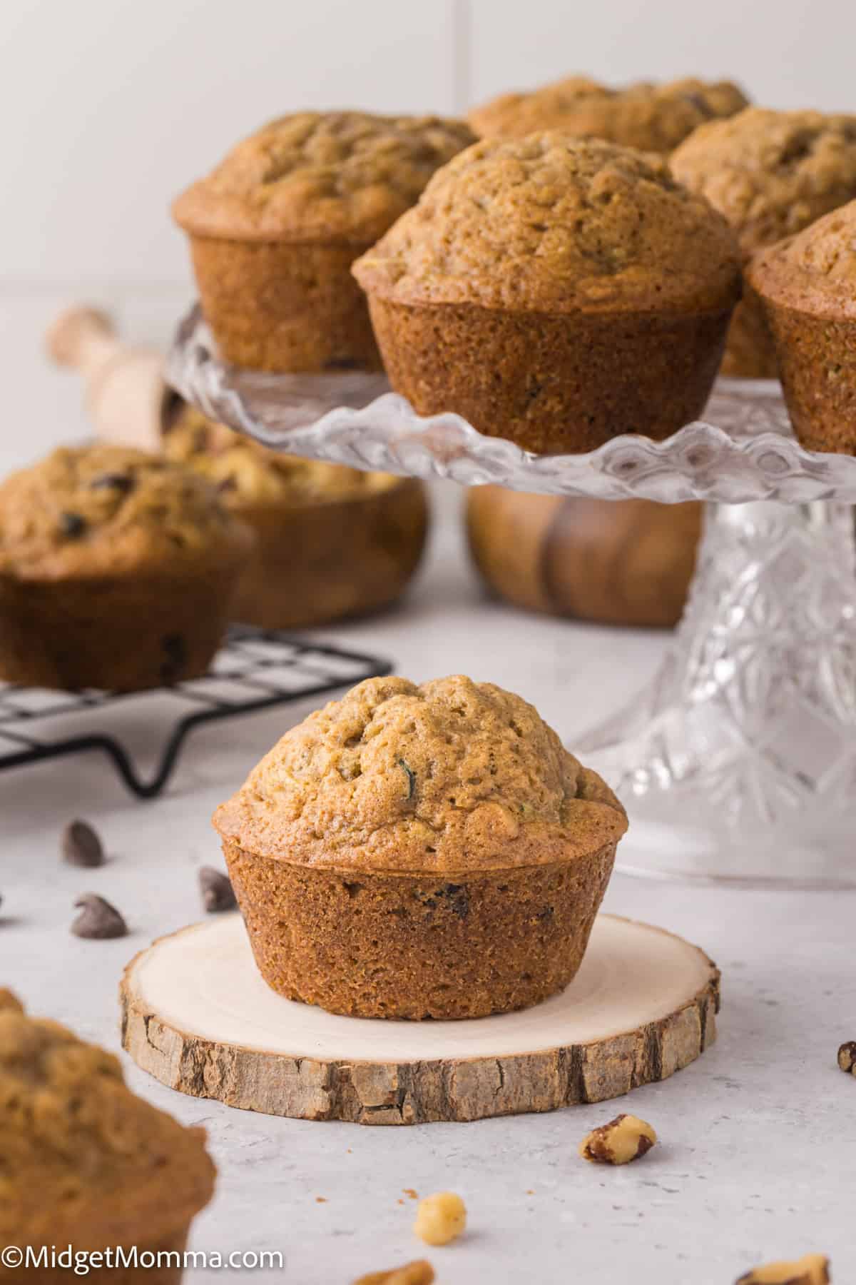 A display of freshly baked muffins on a glass cake stand, with more muffins and chocolate chips scattered on a white surface.